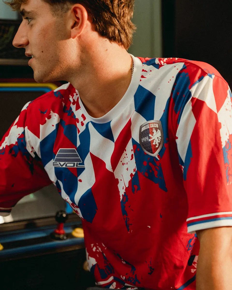 Dettaglio della maglia rossa 2026/27 a motivi geometrici bianchi e blu con stemma e logo Evol