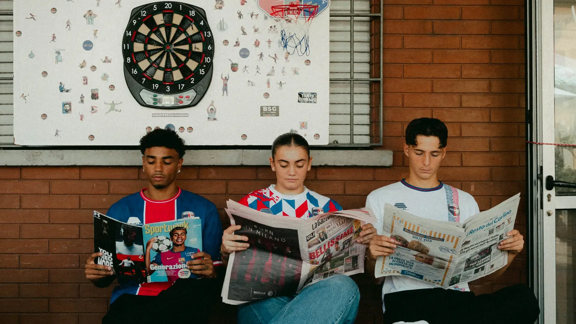 Tre ragazzi con le maglie dell’Imolese leggono il giornale sotto un bersaglio per freccette